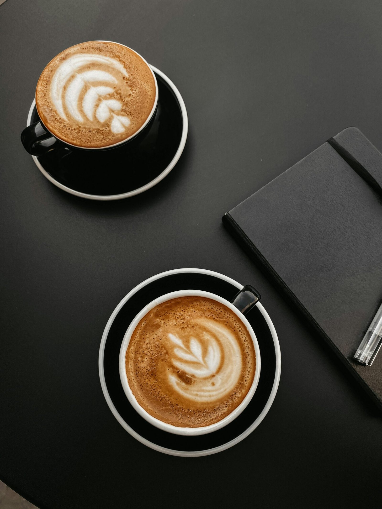 Two beautifully crafted lattes with artistic foam next to a notebook on a sleek black desk.